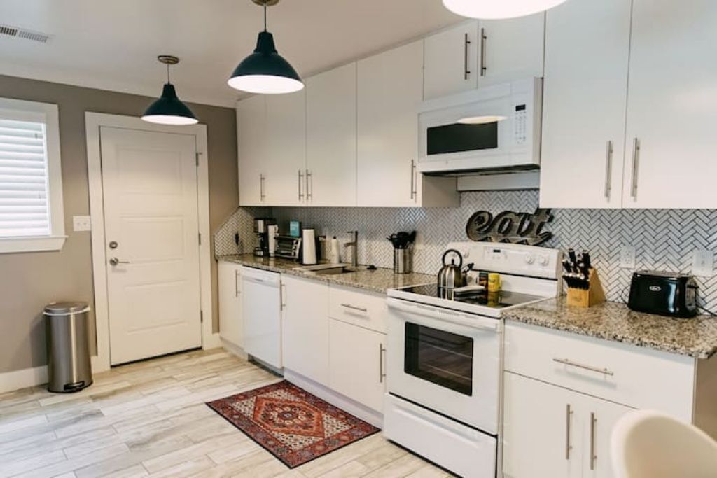 A modern kitchen with white cabinets, a microwave, stove, and various appliances. There is a trash can and a door visible.