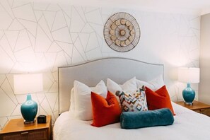 Una cama bien hecha con varios almohadones, dos mesitas de noche con lámparas y un diseño geométrico en la pared.