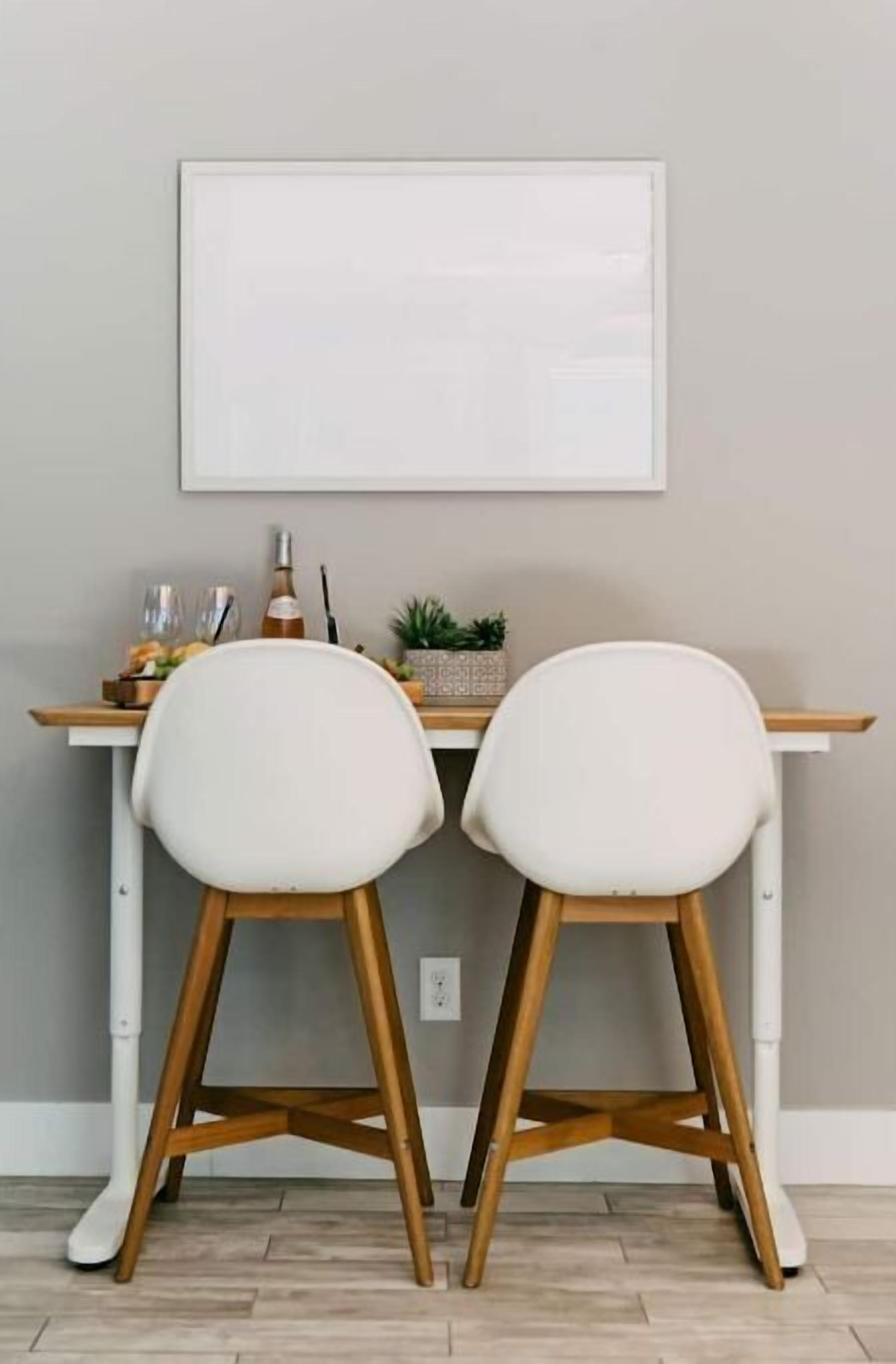 Un comedor con una mesa blanca y dos sillas blancas de patas de madera. Encima de la mesa hay un cuadro enmarcado, dos copas de vino y una botella.