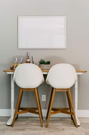 Un comedor con una mesa blanca y dos sillas blancas de patas de madera. Encima de la mesa hay un cuadro enmarcado, dos copas de vino y una botella.