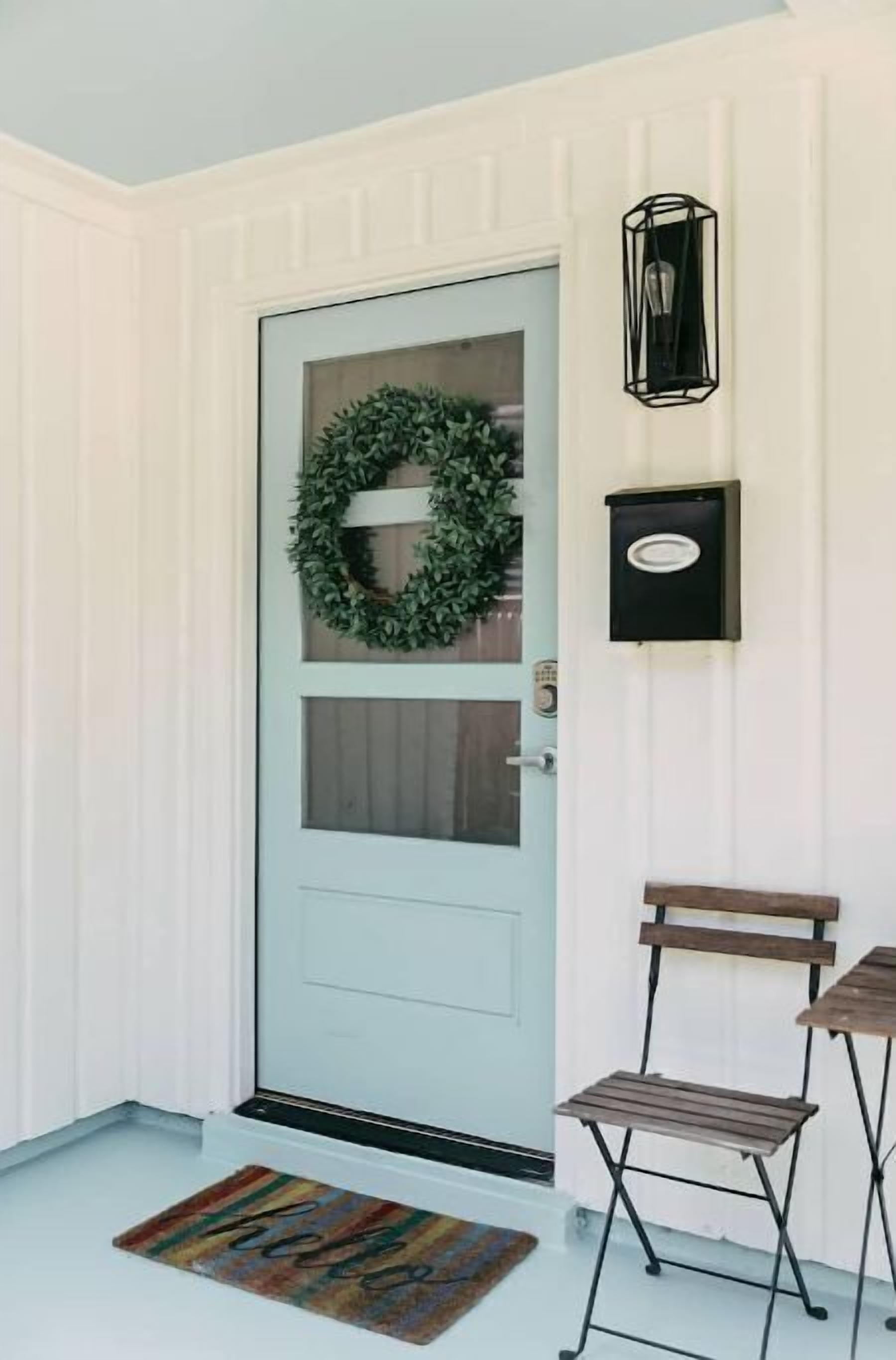 Una puerta blanca con un adorno navideño verde, una luz negra empotrada en la pared y un banco de madera afuera.