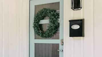 Una puerta blanca con un adorno navideño verde, una luz negra empotrada en la pared y un banco de madera afuera.