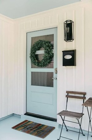 Una puerta blanca con un adorno navideño verde, una luz negra empotrada en la pared y un banco de madera afuera.