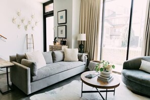 Una sala de estar moderna con un sofá gris, una mesa redonda de centro y vistas a una casa a través de grandes ventanales.