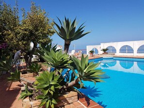 A pool area with a clear blue sky, surrounded by lush greenery and a white building with arched windows. - Villa Velia Priscope (Calabria)