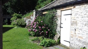Um edifício de pedra com porta branca, rodeado por vegetação exuberante e flores cor-de-rosa.