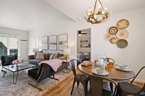 A modern living room with a dining area, featuring a chandelier, artwork, and a coffee table.