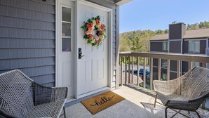 A front porch with a white door, a wreath, two wicker chairs, and a 'hello' mat.