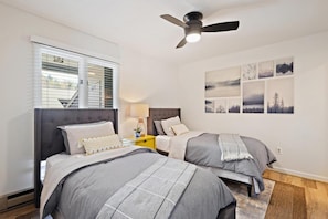 A bedroom with two beds, a ceiling fan, a window with blinds, and artwork on the wall.