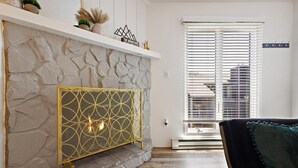 A stone fireplace with a geometric gold radiator, a white mantelpiece, and a window with blinds.