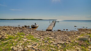A rocky shoreline with a dock extending into calm water, a boat moored at the dock's end, and a clear sky above. - Prairie-Style Home on Garden Bay w/ Deck + Hot Tub (Garden)