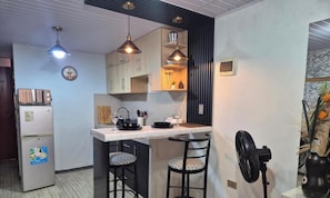 A compact kitchen with a central island, pendant lights, a refrigerator, and a fan.