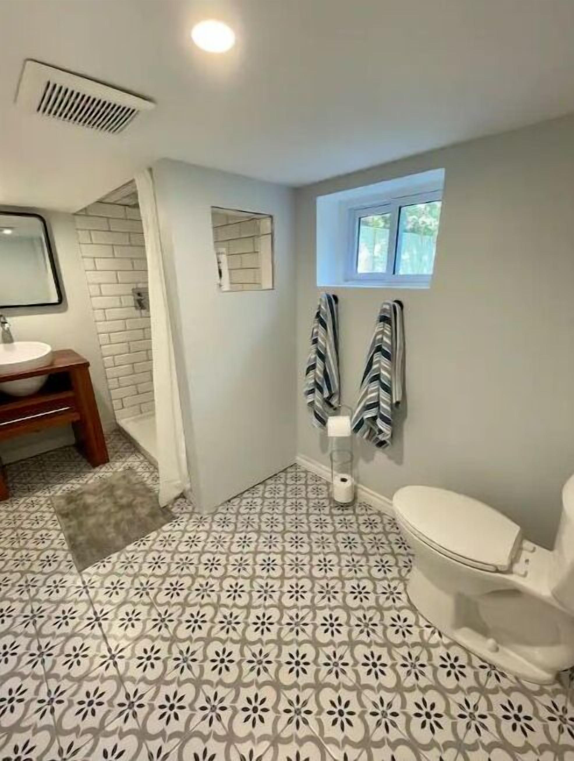 A bathroom with a patterned floor, a toilet, a sink, a mirror, and a window.