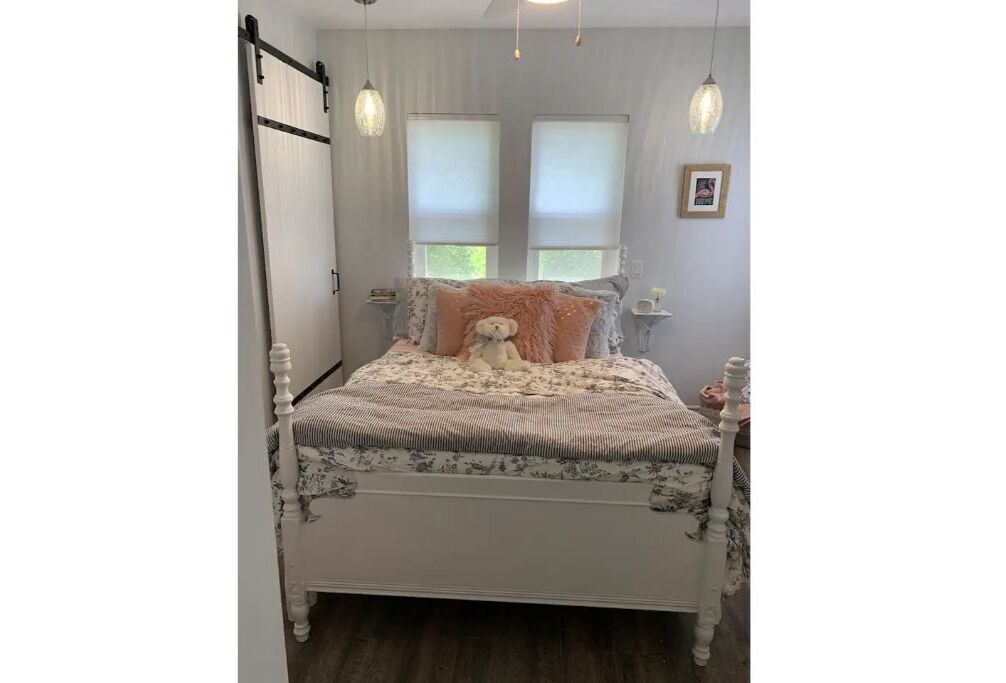 Ein Bett mit floralem Überwurf, einem Teddybären und zwei Kissen. Zwei Fenster mit Jalousien, eine Hängelampe und ein Bild an der Wand sind ebenfalls zu sehen.