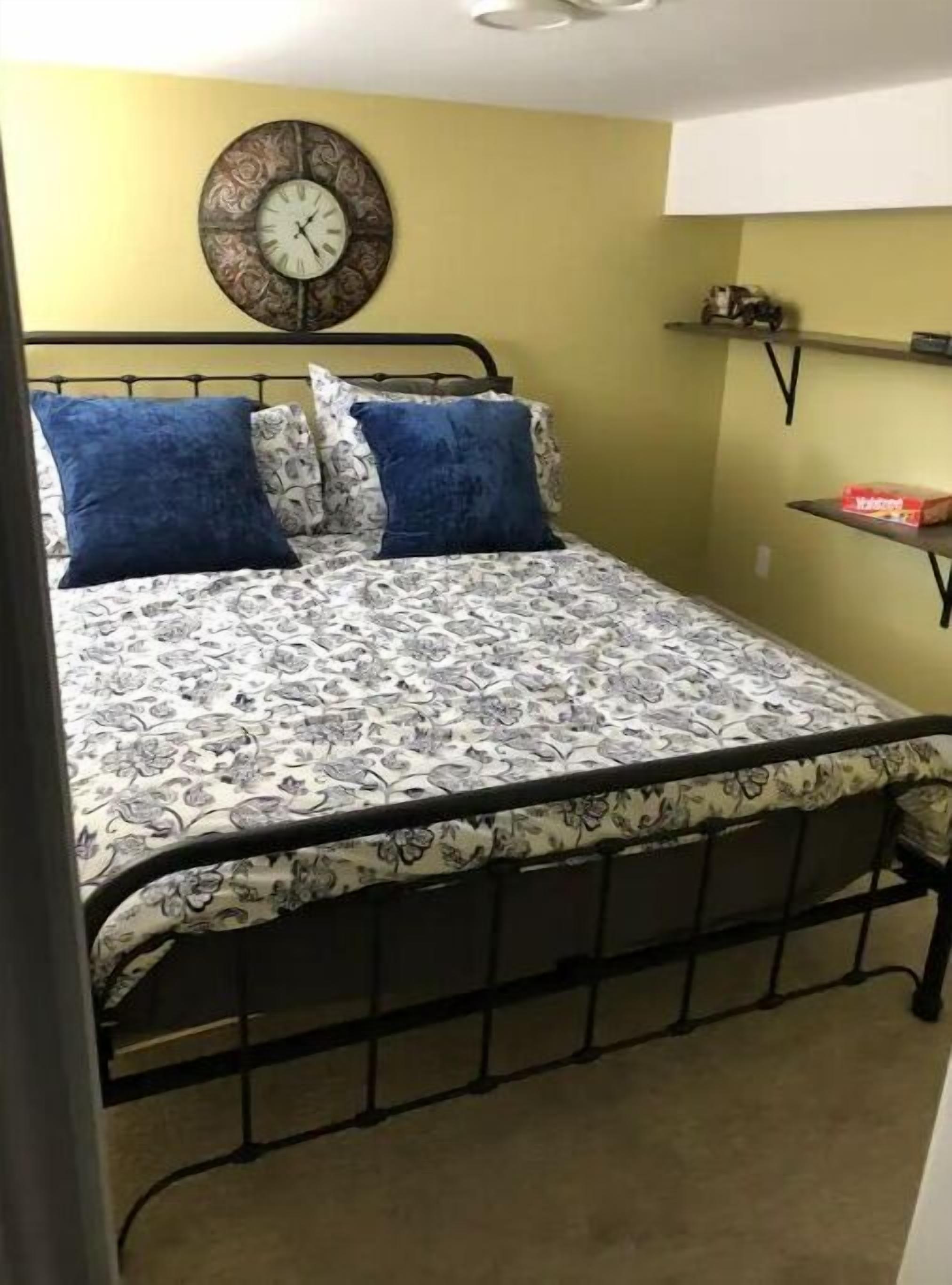 A bedroom with a bed, two blue pillows, a patterned bedspread, a wall clock, and a shelf with a box.