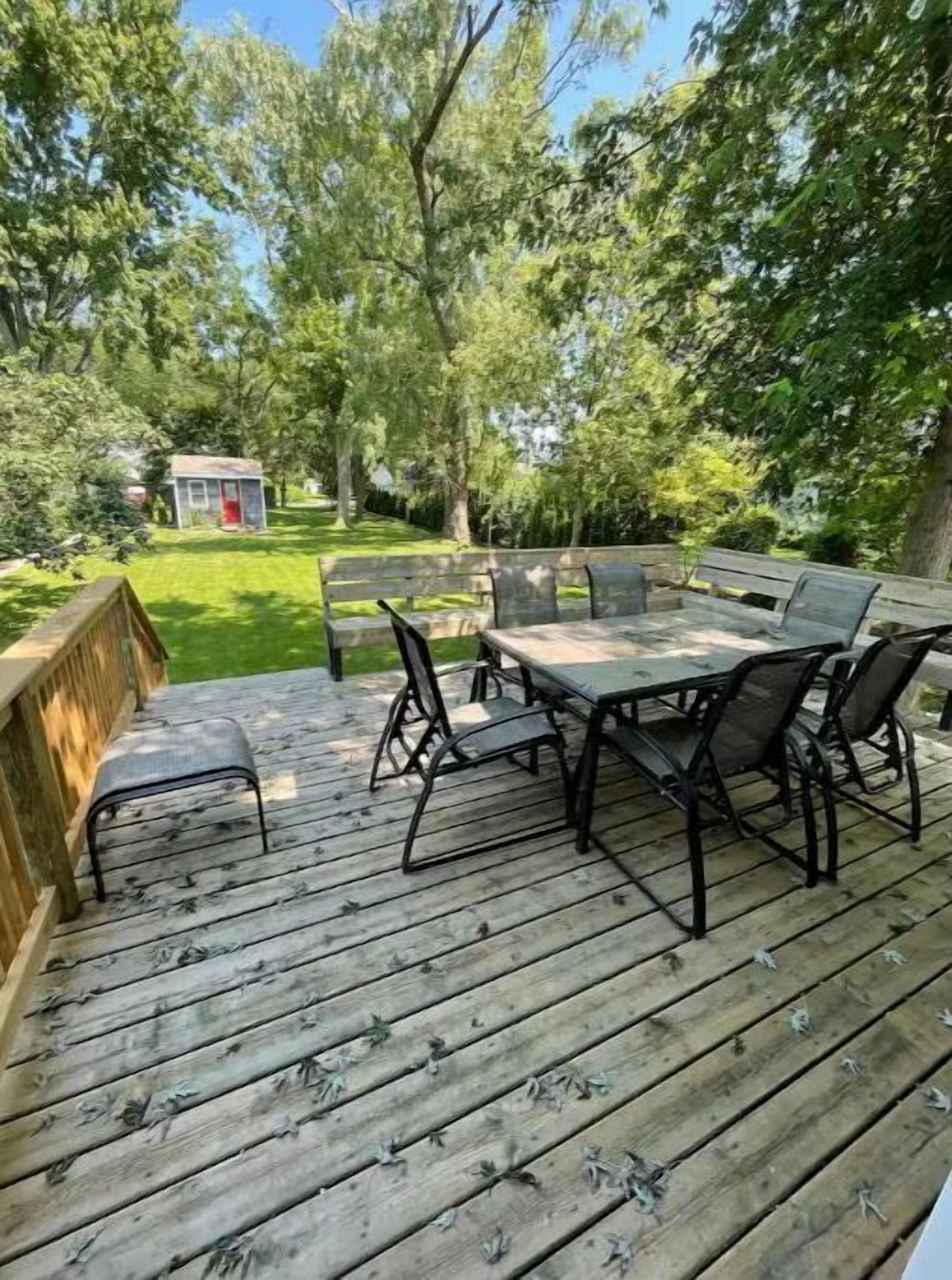Eine Holzterrasse mit Tisch und Stühlen, umgeben von Bäumen und einem kleinen Schuppen.