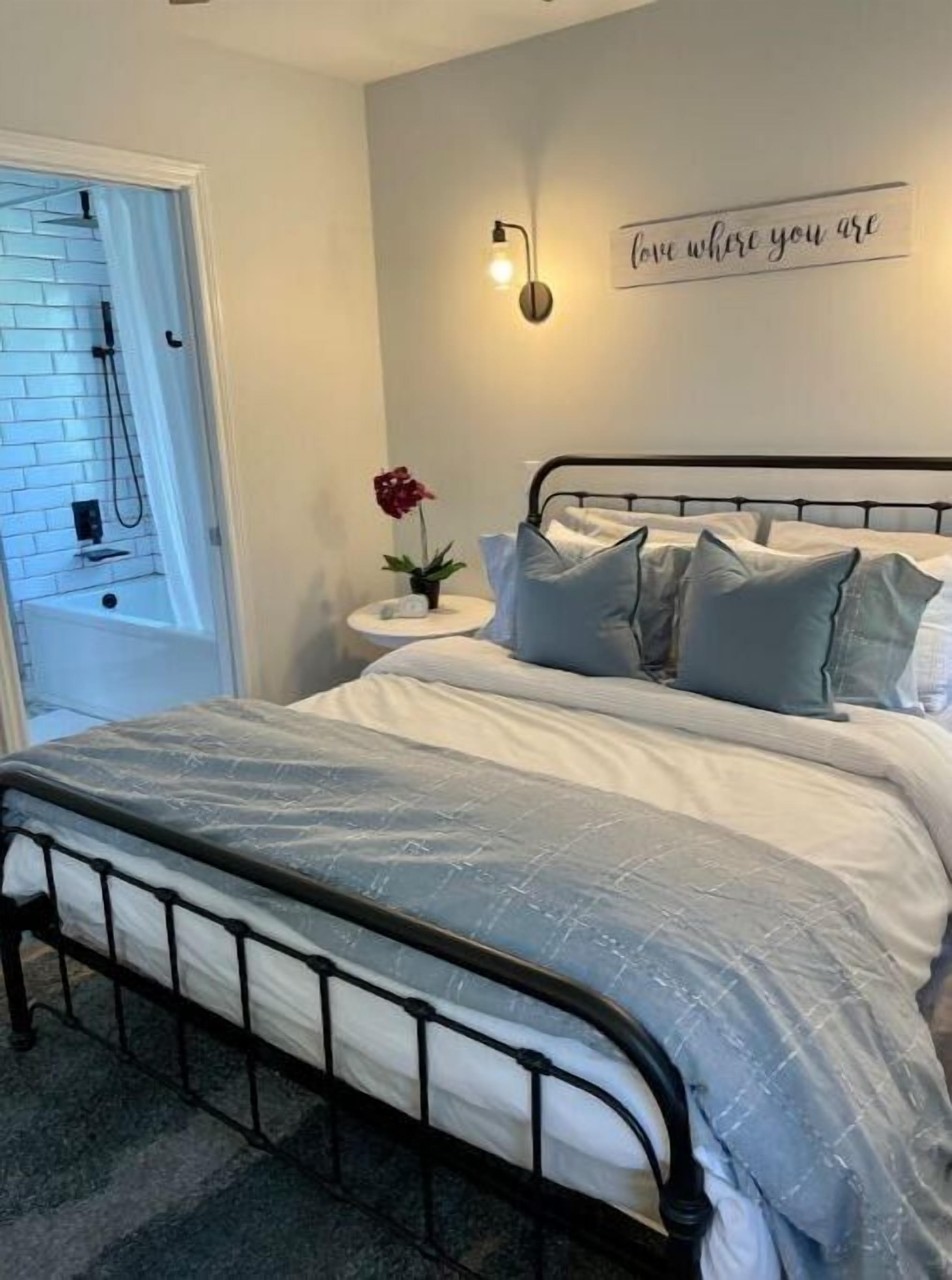 A bedroom with a bed, a nightstand, a wall-mounted light, and a sign that reads "love where you are".