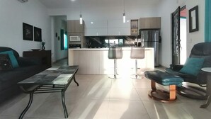 Una sala de estar moderna con un sofá, una mesa de centro y una mesa de comedor. Hay una zona de cocina con un refrigerador, microondas y taburetes de bar.