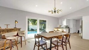A spacious dining area with a wooden table and chairs, a modern chandelier, and a view of a pool through large sliding doors. - Lovely home with heated Pool a mile from the beach (Boca Raton)