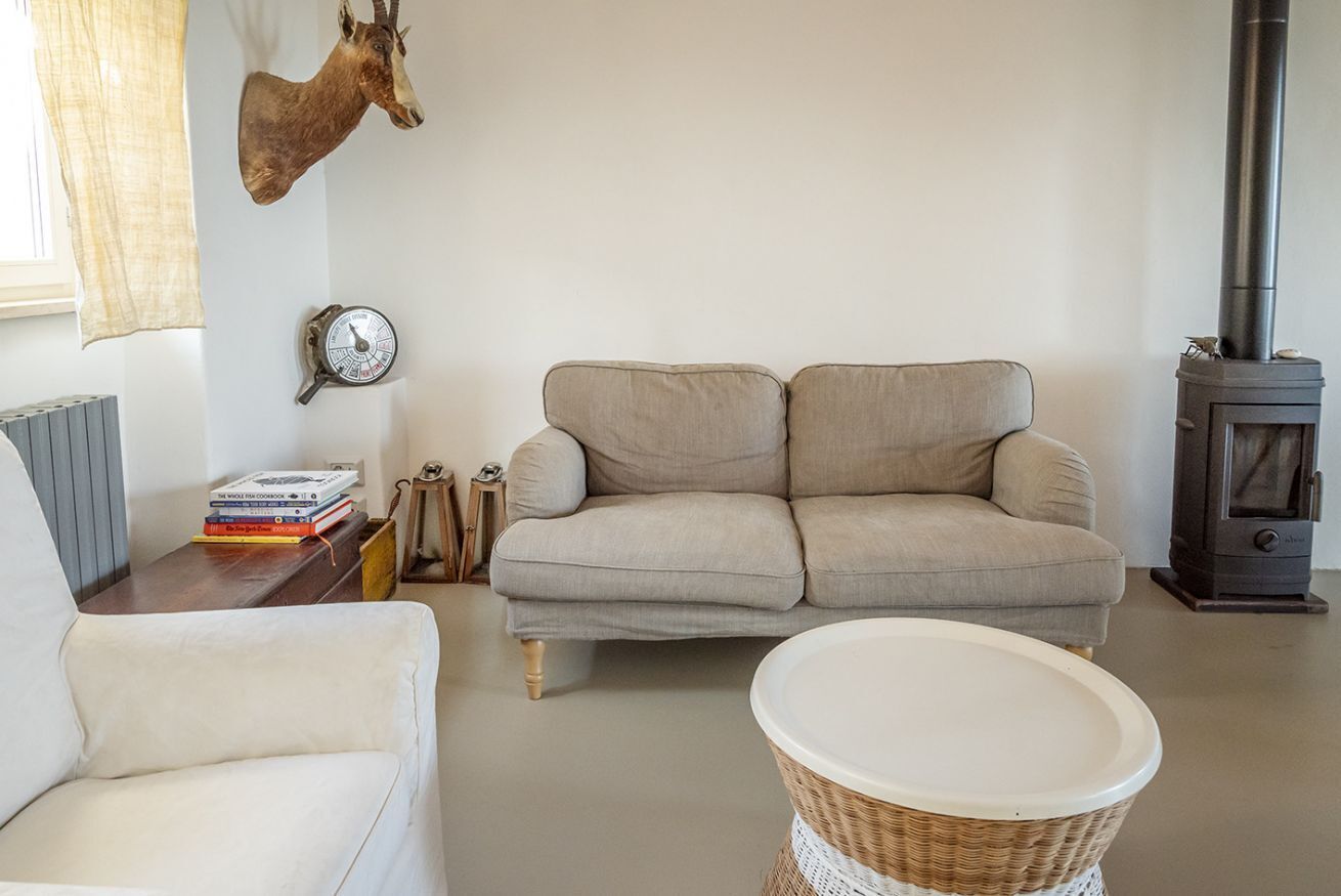 A living room with a sofa, a chair, a wall-mounted clock, a bookshelf, a decorative deer head, and a wood-burning stove.