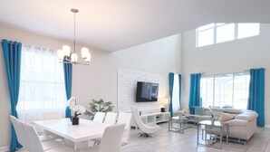 A modern living room with a dining area, white furniture, a flat-screen TV, and large windows with blue curtains. - Superhero Game Room • Mickey Room • Pool & Spa • 4 Miles to Disney (Kissimmee)