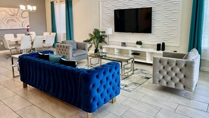 A modern living room with a blue sofa, a grey armchair, a glass coffee table, a white entertainment unit, and a flat-screen TV mounted on a textured wall. - Superhero Game Room • Mickey Room • Pool & Spa • 4 Miles to Disney (Kissimmee)