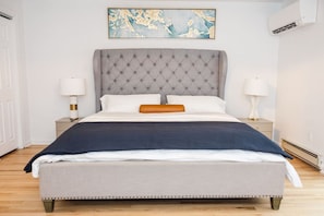 Un dormitorio con cabecera capitonada, cama con ropa de cama blanca y azul oscuro, dos mesitas de noche con lámparas y un cuadro abstracto grande sobre la cama.