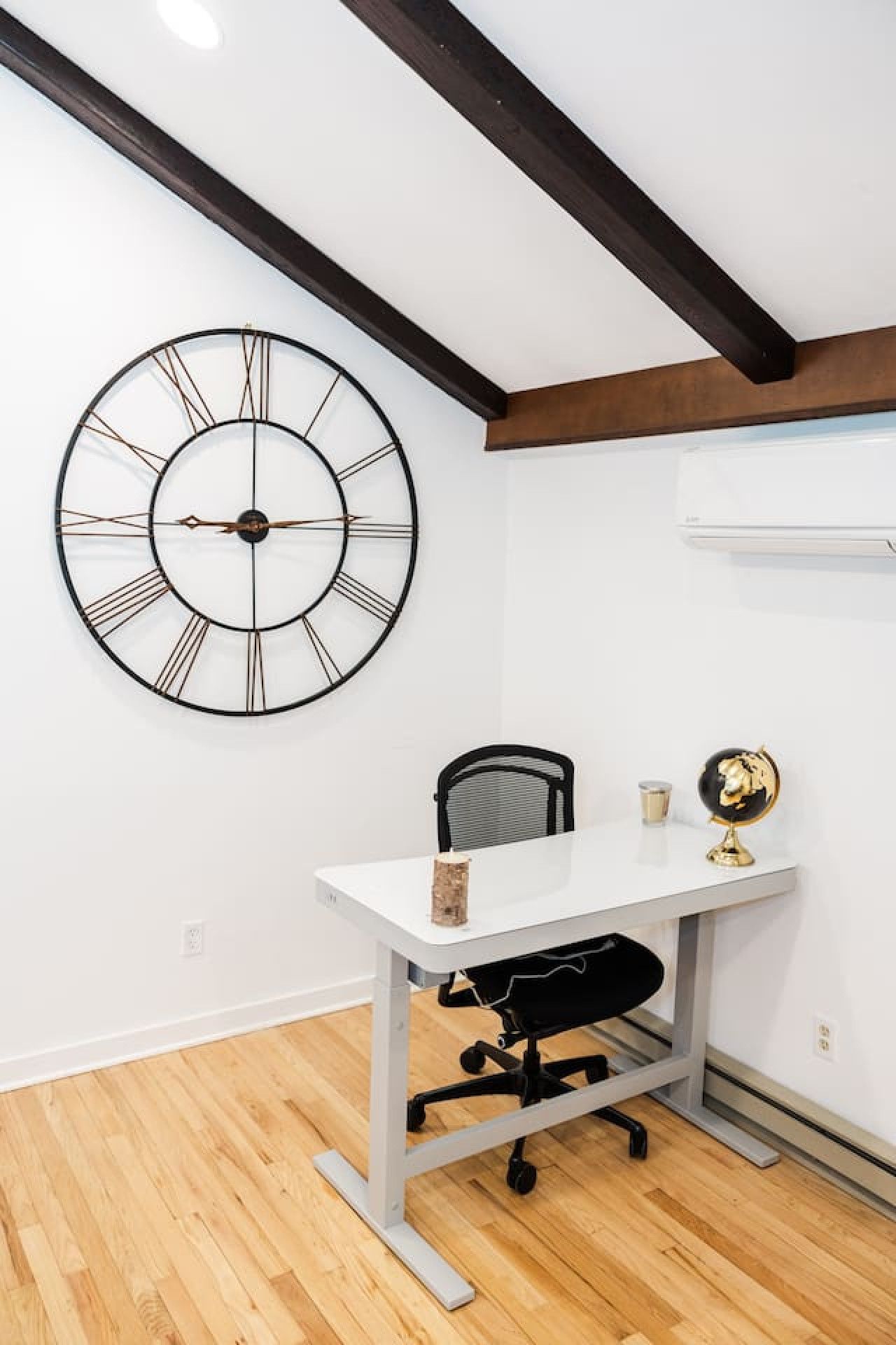 Ein Zimmer mit weißem Schreibtisch, schwarzem Bürostuhl, großer Wanduhr und kleinem Globus an der Wand.