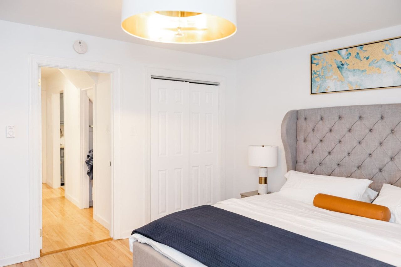 A bedroom with a tufted headboard, a bed with white and dark blue bedding, a white door, and a hallway leading to another room.