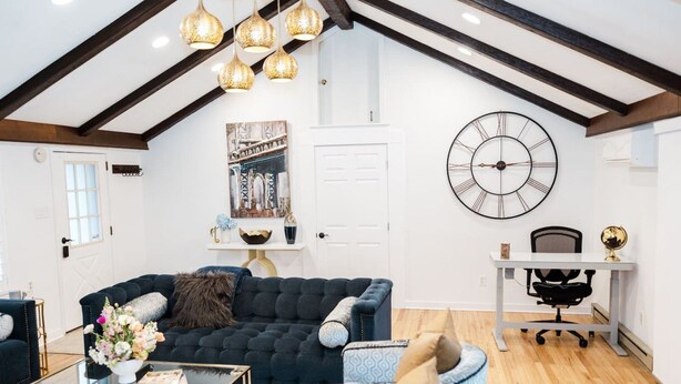 A living room with a dark blue sofa, a glass coffee table, and a white desk with a chair. A large wall clock and a framed picture are also visible. - Cowry Acres - The First House Villa w Hot Tub (New Hope)