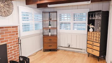 Une pièce avec une cheminée en briques, des murs lambrissés blancs, un plancher en bois, une petite commode en bois, un meuble noir avec des tiroirs et une fenêtre avec des volets blancs.