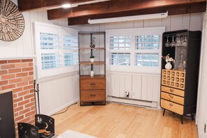 Una habitación con chimenea de ladrillo, paredes blancas con paneles, suelo de madera, un pequeño aparador de madera, un mueble negro con cajones y una ventana con contraventanas blancas.