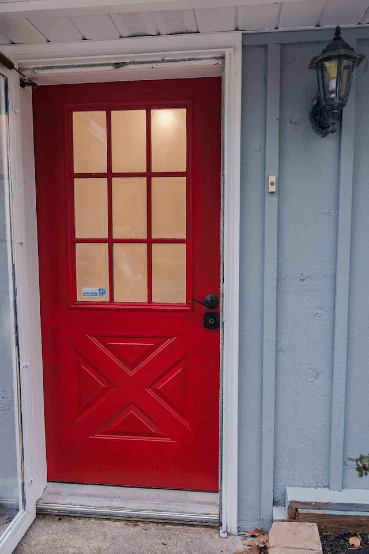 A red door with a glass panel and a geometric design, a black doorbell, and a lantern on the wall.