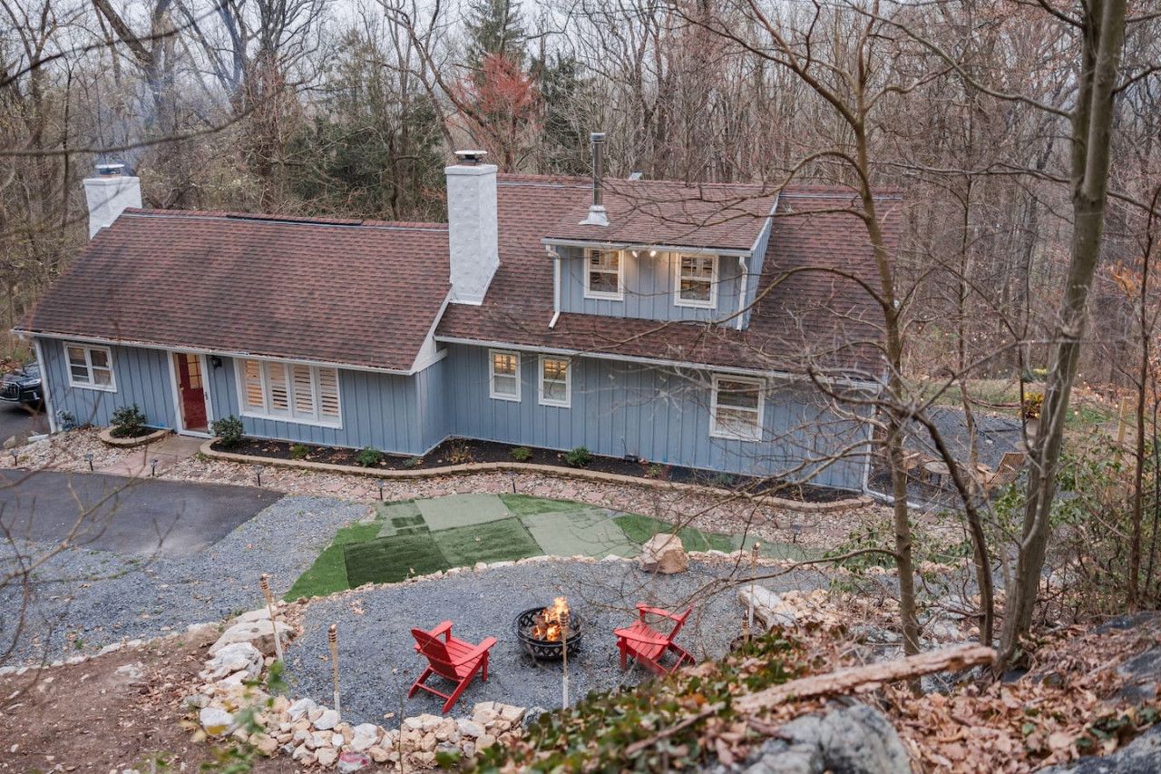 A two-story house with a red roof and blue siding, a front yard with a fire pit and red chairs, and a gravel driveway.