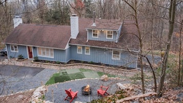 A two-story house with a red roof and blue siding, a front yard with a fire pit and red chairs, and a gravel driveway.