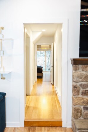A narrow hallway with wooden flooring leading to a room with a bed and a window.