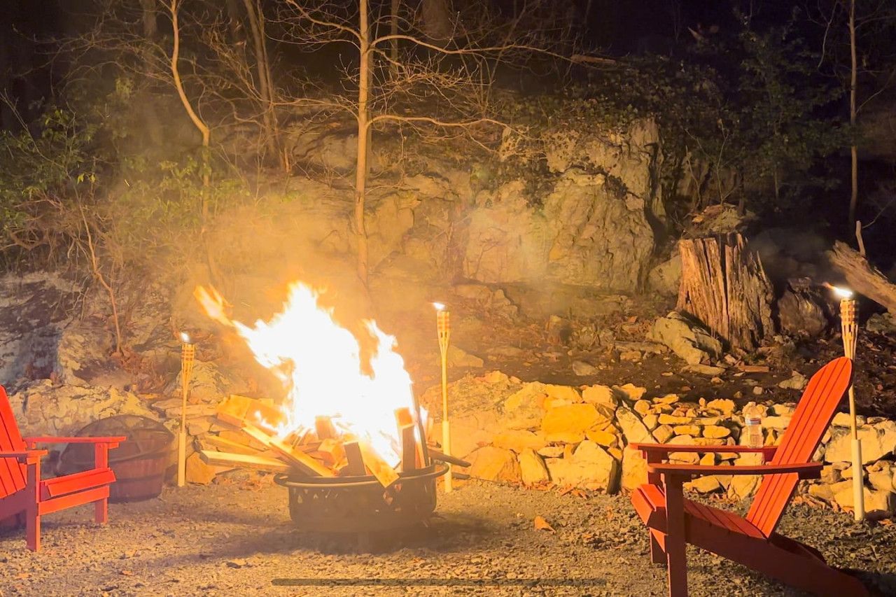 Ein Lagerfeuer mit zwei roten Stühlen und einem Topf im Vordergrund, vor einer felsigen Klippe und Bäumen im Hintergrund.