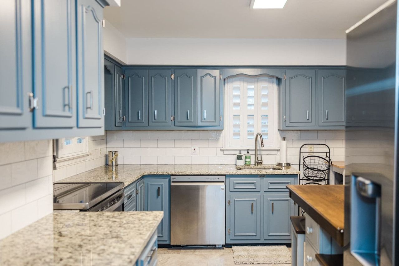 A kitchen with blue cabinets, a stainless steel dishwasher, granite countertops, and a window with blinds.
