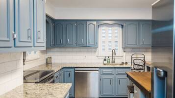 A kitchen with blue cabinets, a stainless steel dishwasher, granite countertops, and a window with blinds.