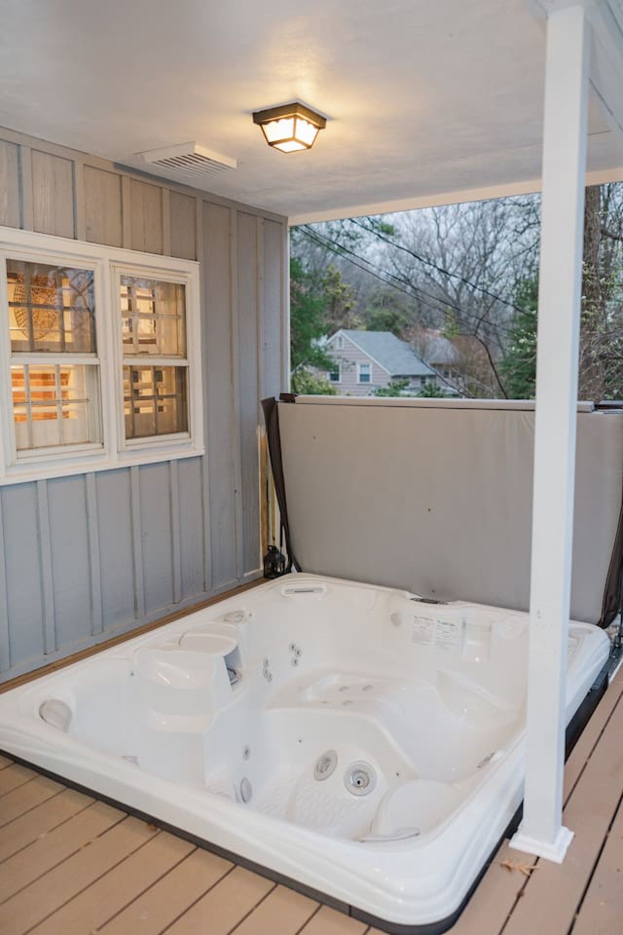 A hot tub on a wooden deck with a covered porch, a window with a view of a house, and a ceiling light fixture.