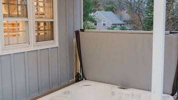 A hot tub on a wooden deck with a covered porch, a window with a view of a house, and a ceiling light fixture.