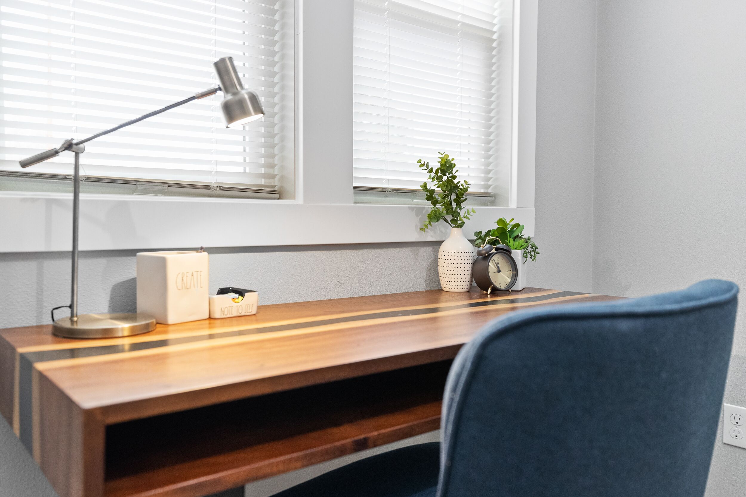 Un escritorio de madera con una lámpara, un jarrón blanco con una planta y un despertador antiguo.
