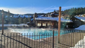 Une zone avec une piscine, délimitée par une clôture en bois, un bâtiment doté d’un toit en pente et, au loin, un paysage enneigé.