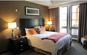 Habitación de hotel con una cama grande, dos mesitas de noche con lámparas, un cuadro enmarcado en la pared y una ventana con cortinas.