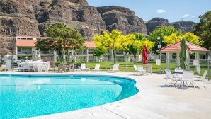 Ein Swimmingpool mit Liegestühlen und Sonnenschirmen, umgeben von Gebäuden und einer Bergkulisse.