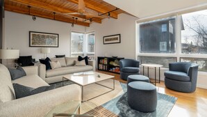 A modern living room with a wooden ceiling, large windows, a sofa set, a coffee table, and a bookshelf.