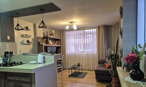 Eine moderne Küche mit Essbereich, einem kleinen Tisch und einer Couch. Ein Fenster mit Vorhängen, ein Deckenventilator und eine dekorative Vase mit Blumen sind ebenfalls vorhanden.