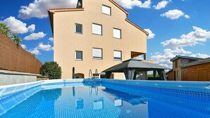Una piscina con baldosas azules cristalinas, una cubierta de madera y un edificio color beige con múltiples ventanas al fondo.