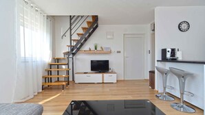 Una sala de estar moderna con una escalera, un televisor sobre un mueble de madera, una cafetera y dos taburetes de bar.