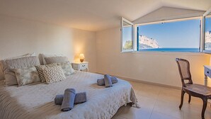Ein Schlafzimmer mit einem großen Bett, einem Sessel, einem Nachttisch und einem Fenster mit Meerblick.